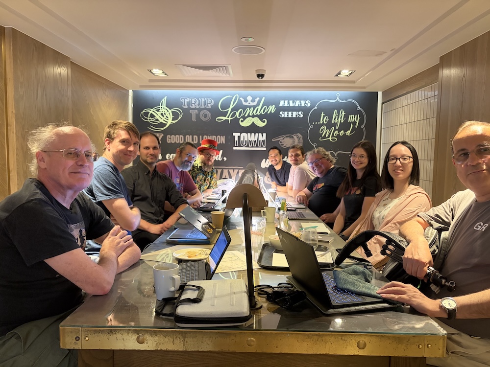 11 attendees sitting round a long table in a caf&eacute; with laptops and the backdrop of a mural-style decorated wall with Trip to London Town