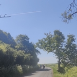 A quiet countryside road with a bunch of cyclists in the distance and jet stream in the clear blue sky above