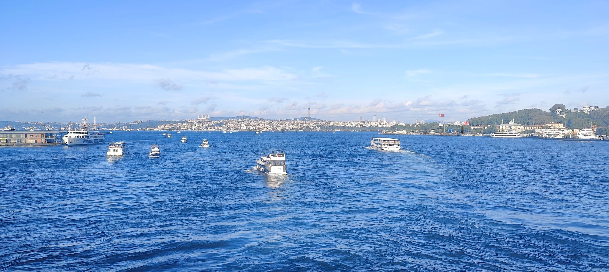 Boats cross the blue waters of the Bosphorus in Istanbul under a clear sky, with the city skyline and Turkish flags visible in the distance.