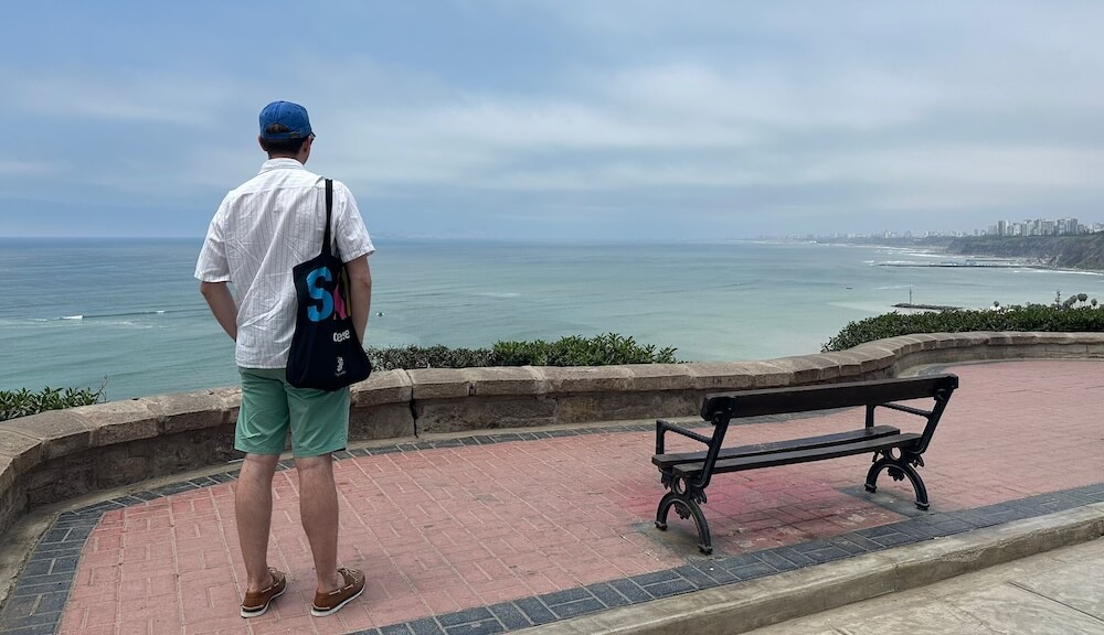 Calum overlooks ocean from Barranco viewpoint, with bench, skyline, and cloudy sky in view.