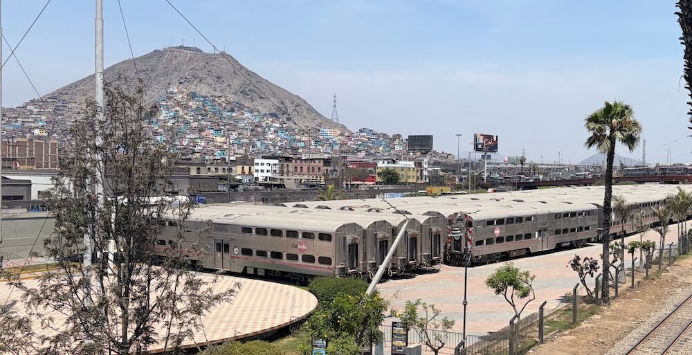 Silver CalTrain cars in yard with hillside shanty homes and mountain backdrop in Lima.