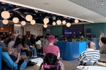 A large audience for GAAD 15 in a colourful office lounge with a range of people in attendance with wheelchairs, and Jamie in their custom made chair