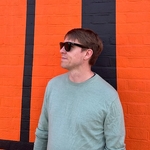 Calum in light green jumper and sun glasses in front of a stricking orange and black painted brick wall
