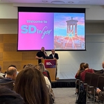 2 men stand preparing to present behind a lecturn with a large screen including the text Welcome to SDinGov and photo Edinburgh's iconic monument with sun rays shining through it