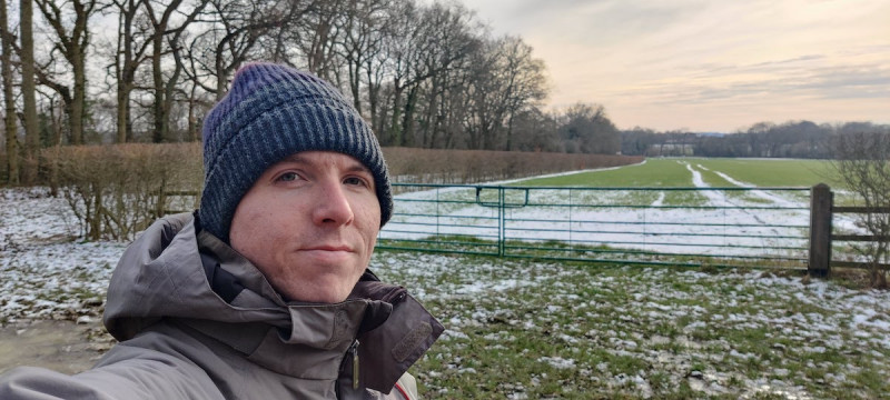 Calum beside a green gate in tree-lined countryside with some snow on the field
