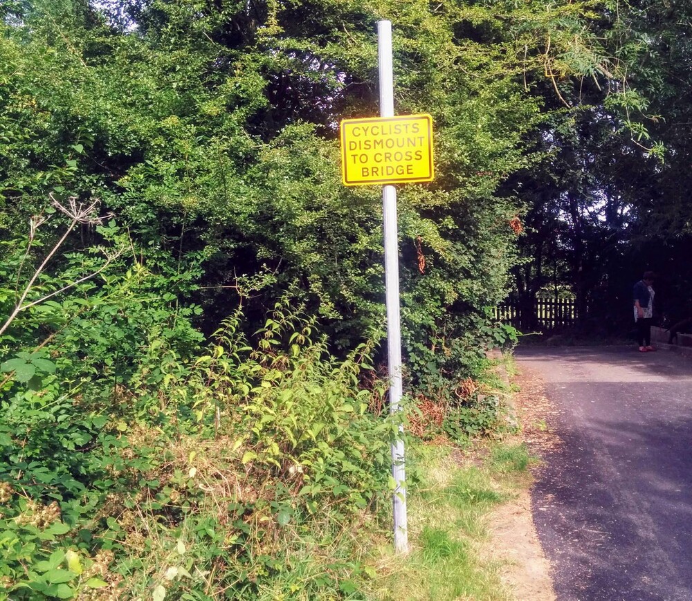 Yellow signpost - cyclists dismount to cross bridge on roadside
