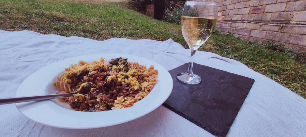 Plate of lentil Bolognese on a white picnic cover on grass beside a glass of white wine