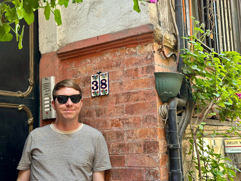 The house number 38 on ceramic tiles with a pretty backdrop of plants and brick wall, Calum stand beside wearing sunglasses