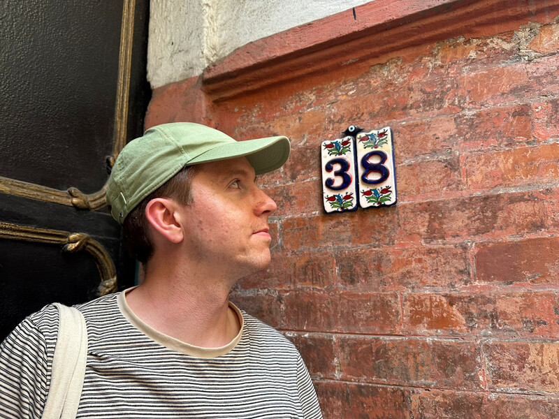 The house number 38 on ceramic tiles with a pretty backdrop of plants and brick wall, Calum stands beside looking at the number
