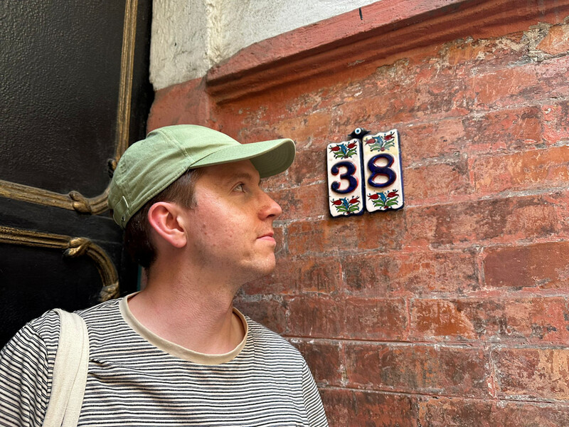 The house number 38 on ceramic tiles with a pretty backdrop of plants and brick wall, Calum stands beside looking at the number