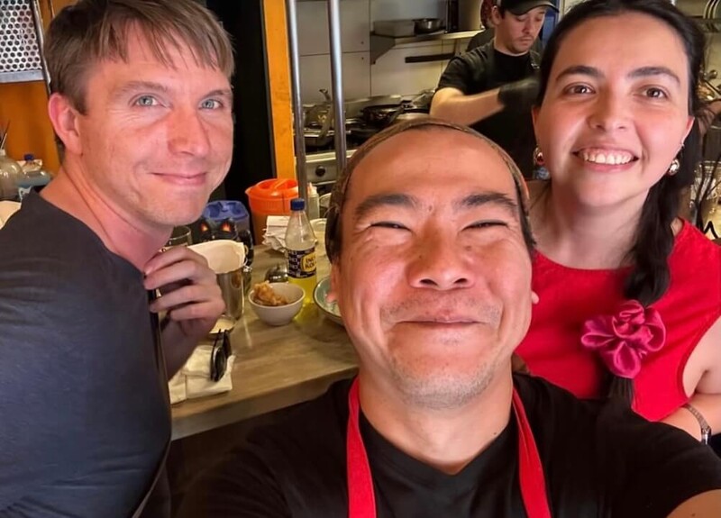Calum and Mena smiling alongside the owner of a restuarant in a selfie pose